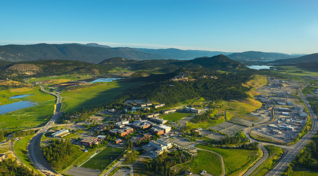 a birds eye view kelowna in a valley surrounded by mountains and the lake in the background, with kelowna's university of british columbia okanagan campus in the centre