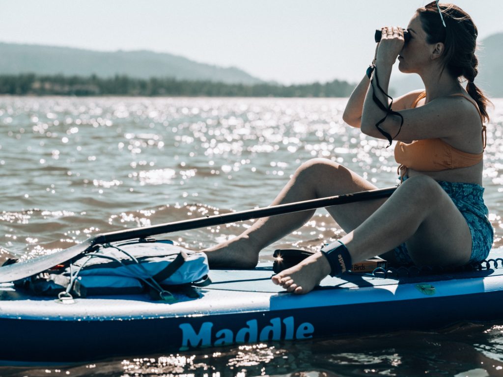 a woman sitting on a stand up paddle board rented from a sporting goods store in kelowna, with binoculars looking over the water
